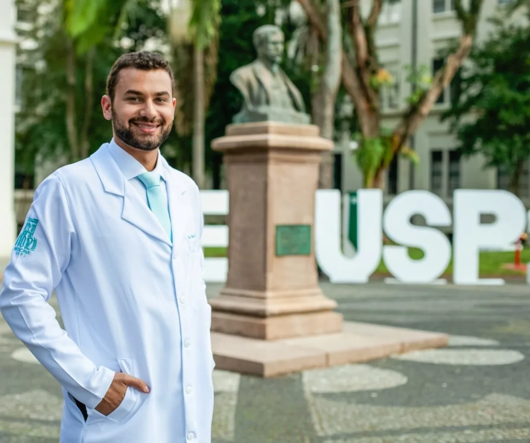Dr. César de jaleco no pátio da Faculdade de Medicina da USP, com o letreiro USP ao fundo
