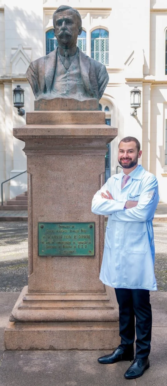 Dr. César de jaleco e braços cruzados ao lado de um busto histórico da Faculdade de Medicina da USP